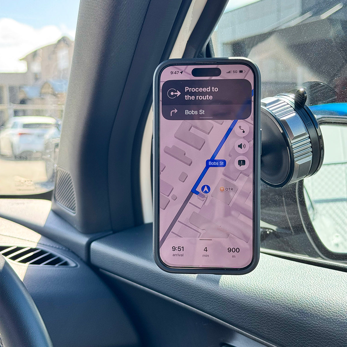 A black magnetic phone mount attached to a car window mirror, holding a smartphone displaying a navigation map. The mount is shown installed in a real car interior.